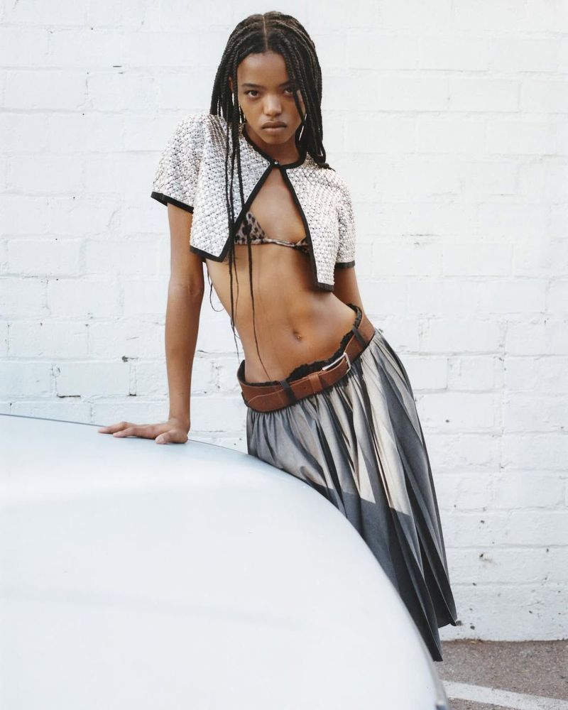 Model leaning on a car wearing a white textured cropped cardigan and pleated skirt against a white brick wall