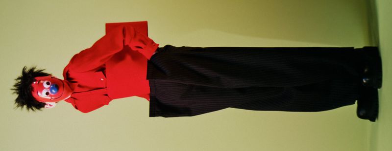 Red puppet with a blue nose and black hair is wearing a red button-down shirt and black pants against a light green background.