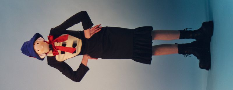 Person in costume with blue hat and red bow poses in front of a light blue wall, wearing black dress and boots with hands on hips.