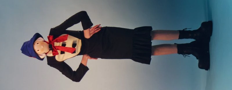 Person in costume with blue hat and red bow poses in front of a light blue wall, wearing black dress and boots with hands on hips.