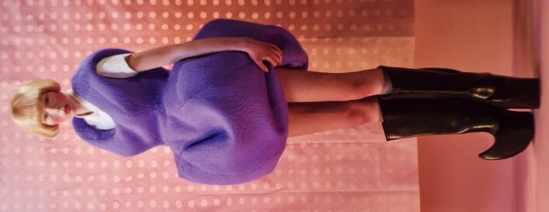 Model wearing a structured purple dress and black boots against a dotted pink backdrop