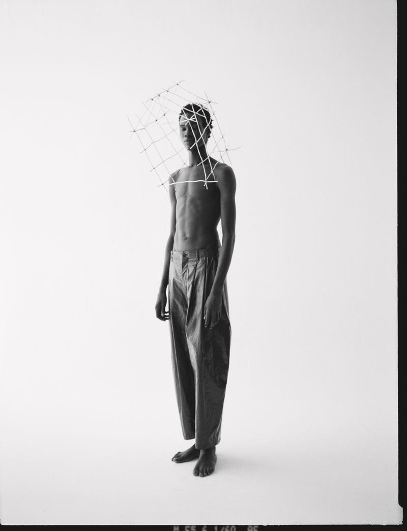 Black and white studio shot of slender, bare-chested person wearing dark pants and an open-frame wire sculpture on their head.