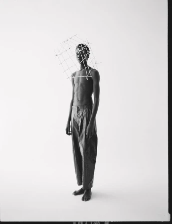 Black and white studio shot of slender, bare-chested person wearing dark pants and an open-frame wire sculpture on their head.