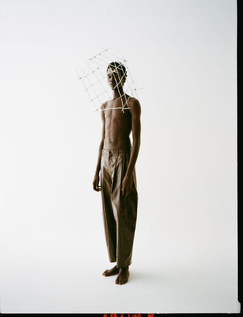 Adult male model in tan trousers with a cage-like headpiece against a white studio backdrop. Fashion editorial style, full-length shot.