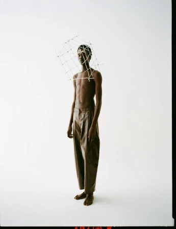 Adult male model in tan trousers with a cage-like headpiece against a white studio backdrop. Fashion editorial style, full-length shot.