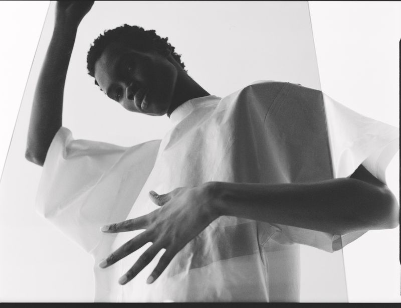 Monochromatic fashion shot of a Black model wearing a flowing white top, striking a dynamic pose against a bright, clean backdrop.