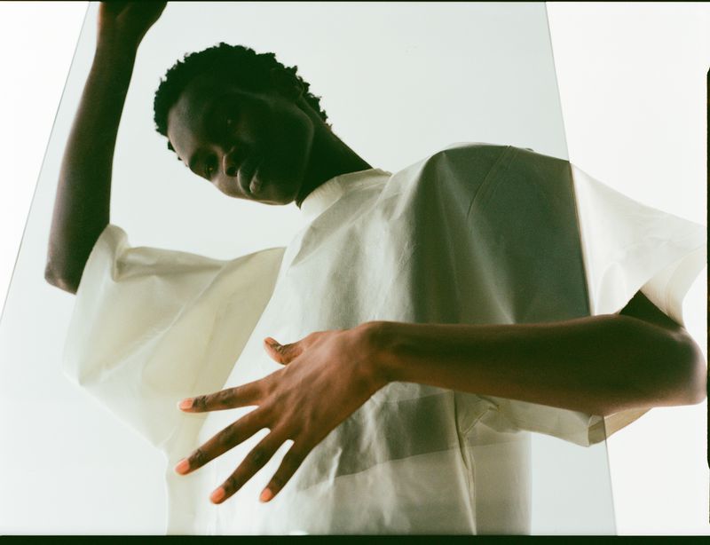Portrait of a young Black woman wearing a white shirt posing behind a clear glass panel, with a neutral expression and soft lighting.