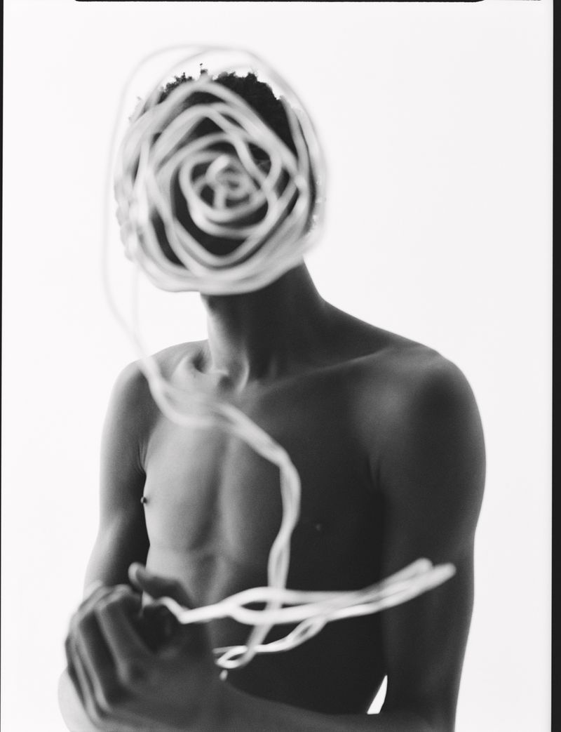 Monochrome portrait of a shirtless person with spiraling white cords obscuring the face against a stark white background.