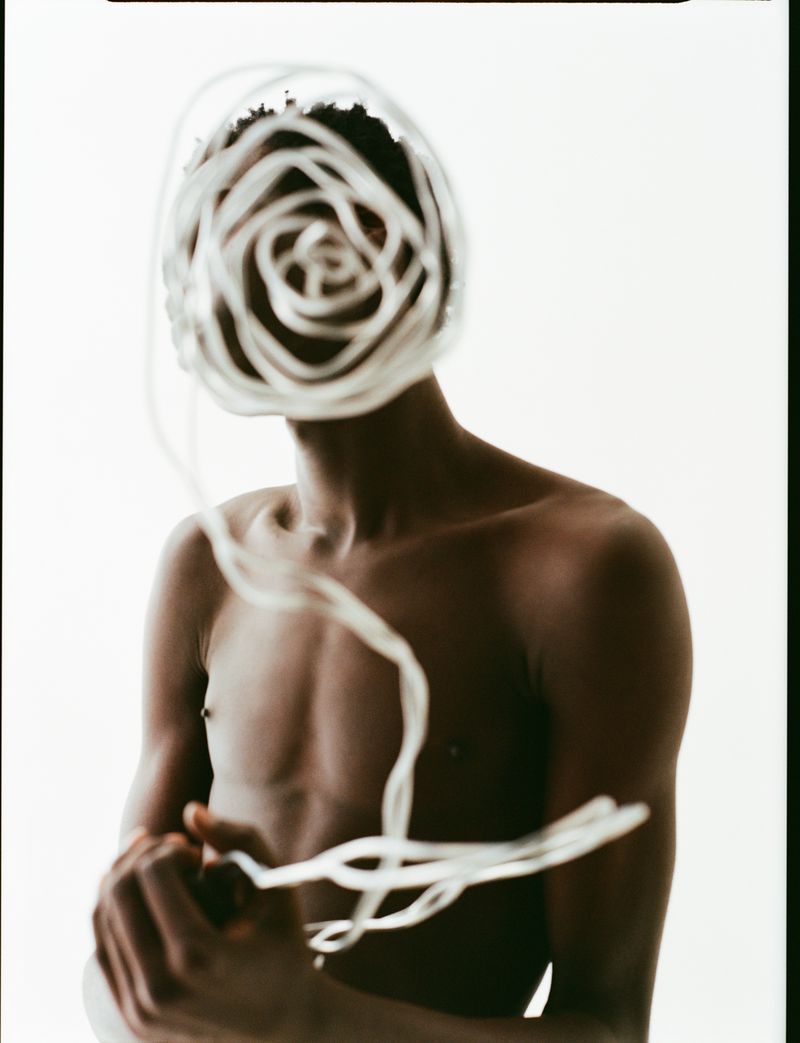 Artistic studio shot of a shirtless Black male model obscured by coiled white cables against a bright, neutral background.