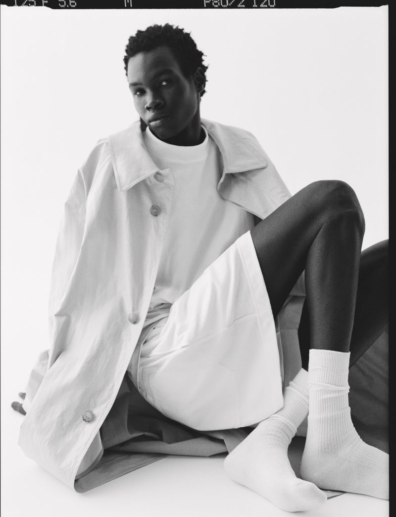 Black model poses in neutral-toned coat and shorts against white background, wearing white socks and a t-shirt, studio lighting.