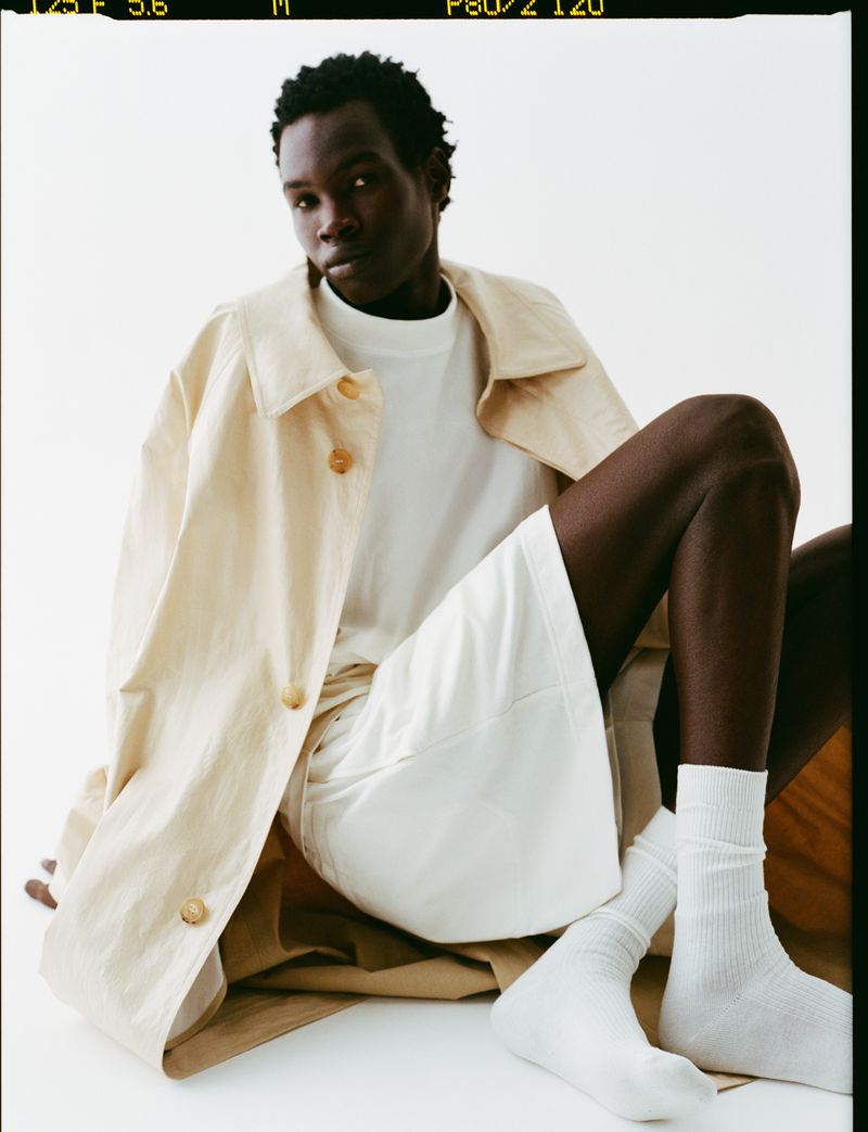 An adult Black male poses in a photo studio wearing a beige button-down coat, white t-shirt, shorts, and socks against a white backdrop.