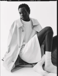 Model seated on the floor wearing a white t-shirt, shorts, white socks, and a light-coloured coat against a plain white backdrop.