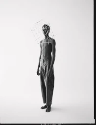Black man standing shirtless wearing trousers with a wire grid structure framing his head against a white background