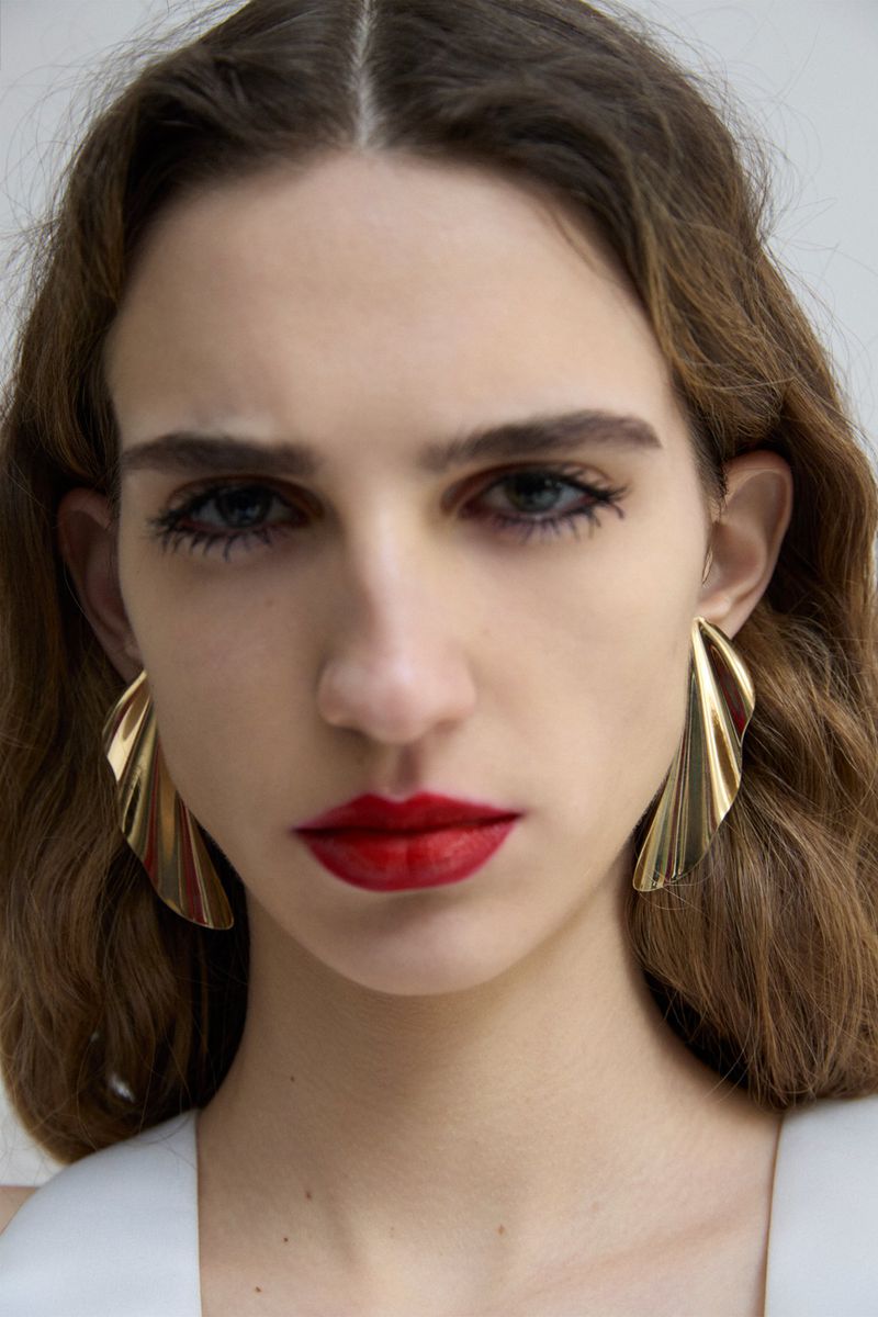 Close-up portrait of a woman wearing statement gold earrings and bold red lipstick against a plain white background.