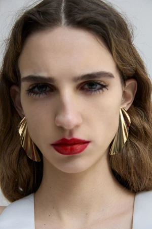 Close-up portrait of a woman wearing statement gold earrings and bold red lipstick against a plain white background.