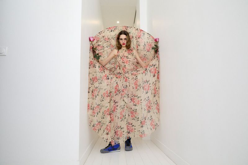 A woman in a floral dress stands in a white hallway, holding roses, with a neutral expression and wearing blue sneakers for a fashion editorial.