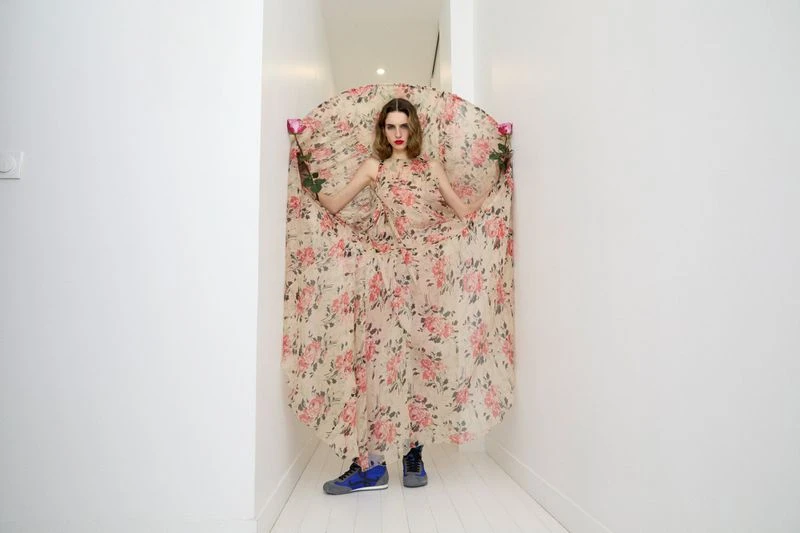 A woman in a floral dress stands in a white hallway, holding roses, with a neutral expression and wearing blue sneakers for a fashion editorial.