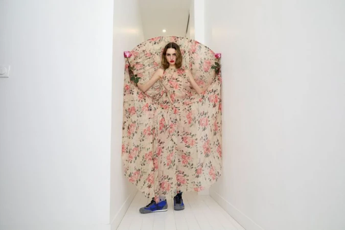 A woman in a floral dress stands in a white hallway, holding roses, with a neutral expression and wearing blue sneakers for a fashion editorial.