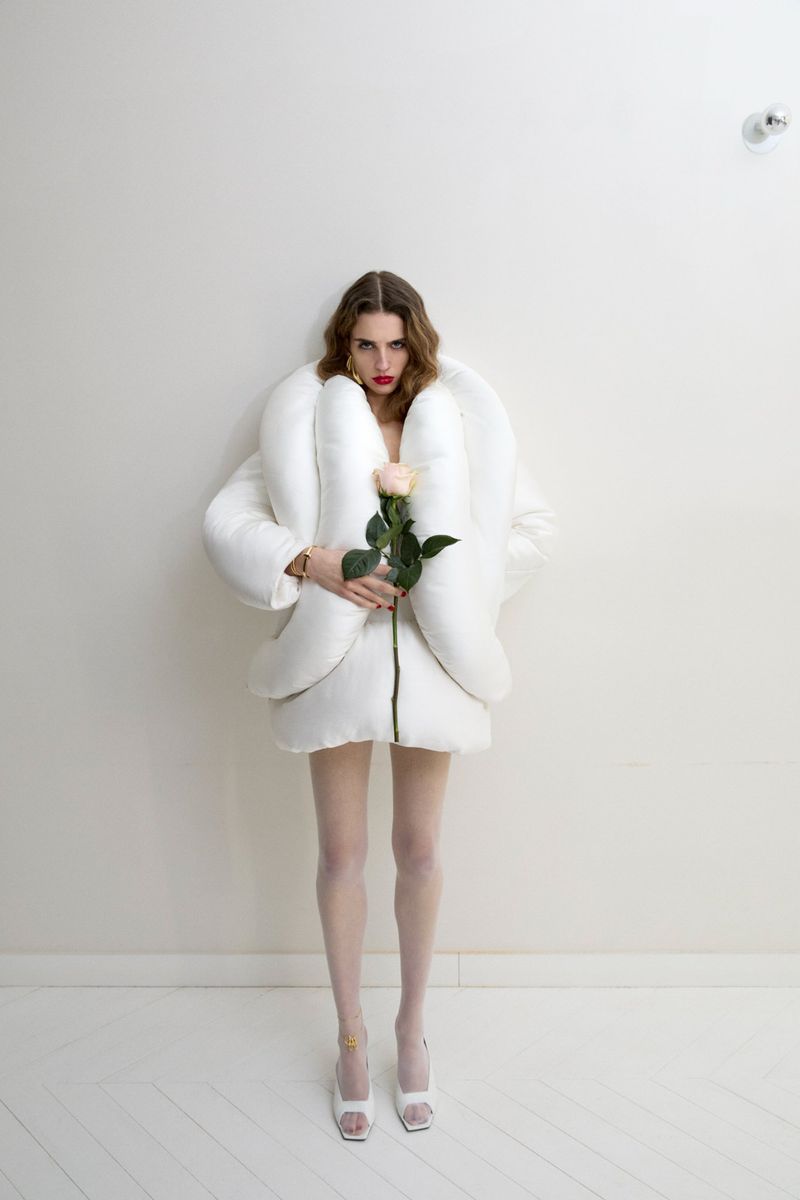 Full shot of a woman in a white puffy jacket holding a peach rose, posing in front of a white wall, and wearing white tights and high heels.