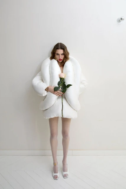 Full shot of a woman in a white puffy jacket holding a peach rose, posing in front of a white wall, and wearing white tights and high heels.