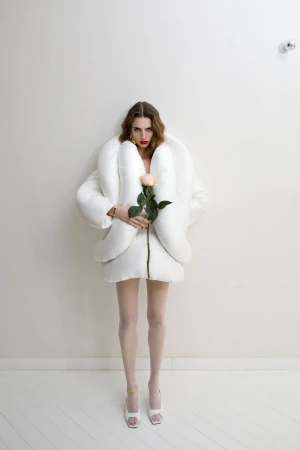 Full shot of a woman in a white puffy jacket holding a peach rose, posing in front of a white wall, and wearing white tights and high heels.