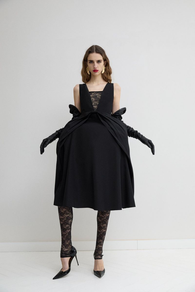 Model in a black sculptural dress with lace tights and long gloves against a plain white studio backdrop