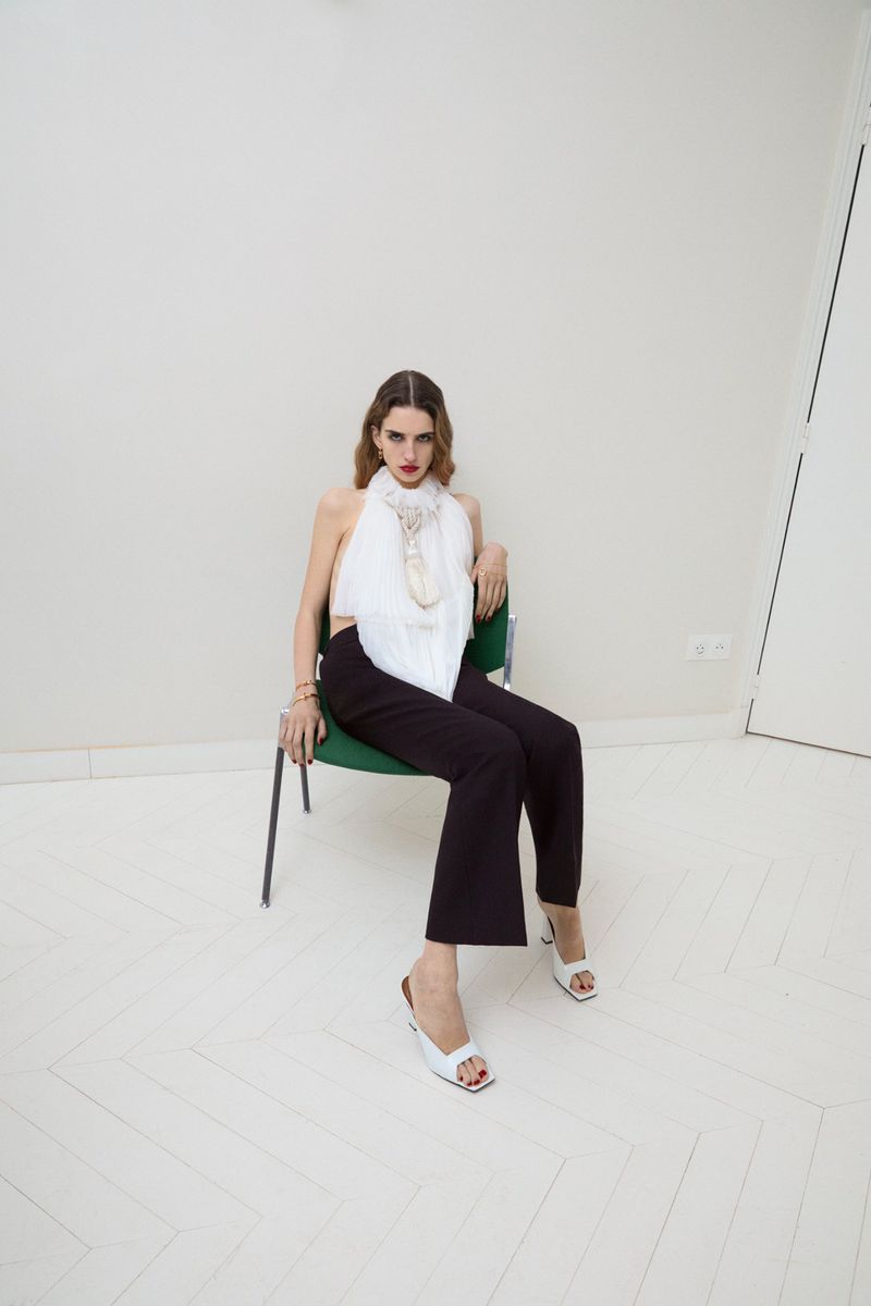 Fashion shot of a woman in a white halter top and black pants sitting on a green chair in a photo studio with a neutral background.