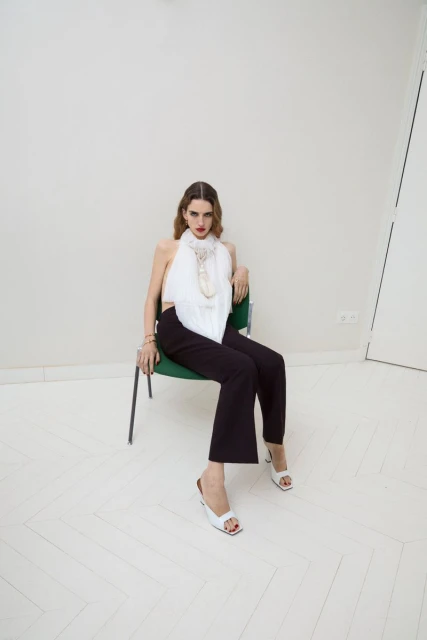 Fashion shot of a woman in a white halter top and black pants sitting on a green chair in a photo studio with a neutral background.