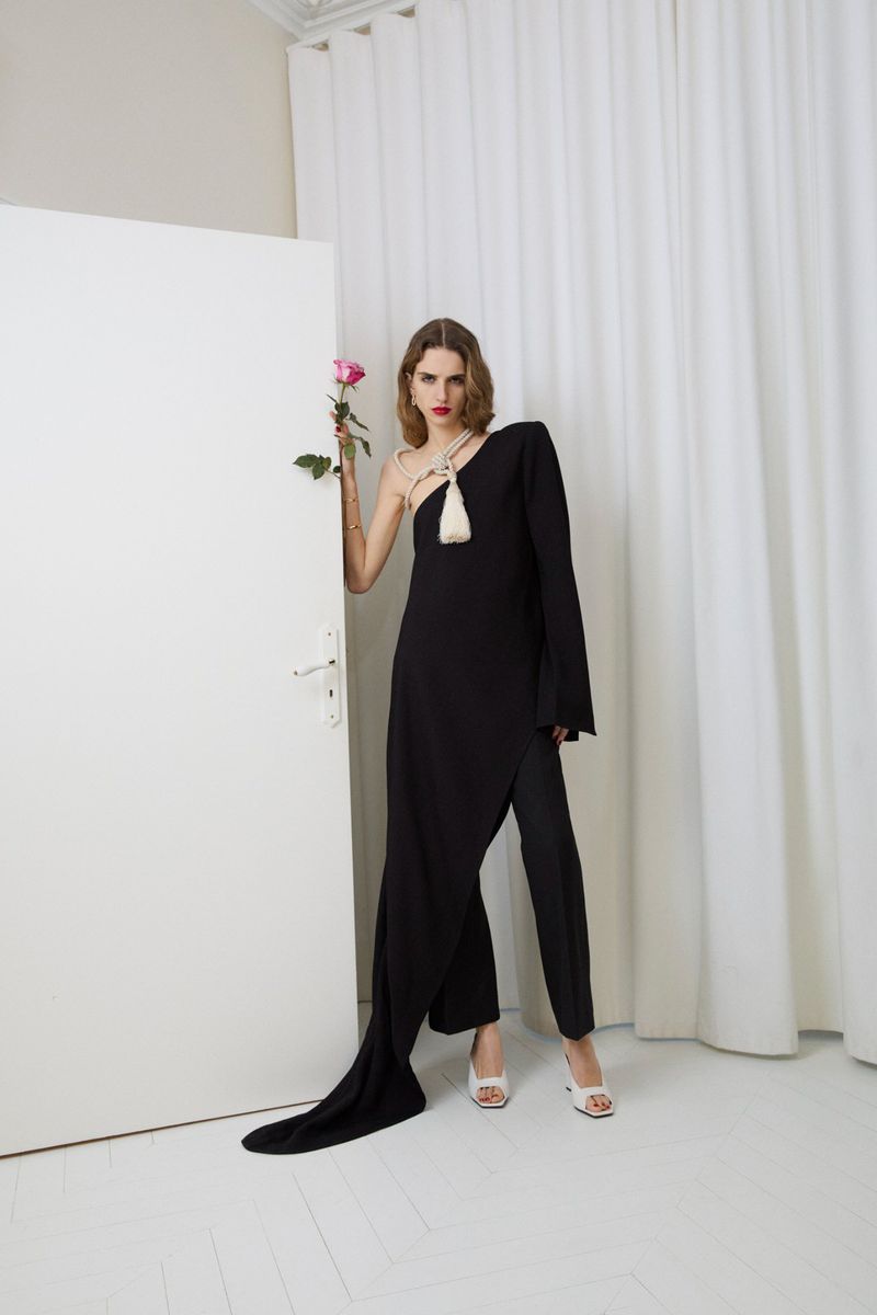 Fashionable woman in black dress and pants, white heels, and holding a pink rose, poses in front of a white door and curtain.