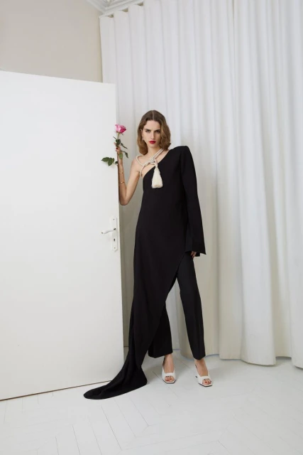 Fashionable woman in black dress and pants, white heels, and holding a pink rose, poses in front of a white door and curtain.