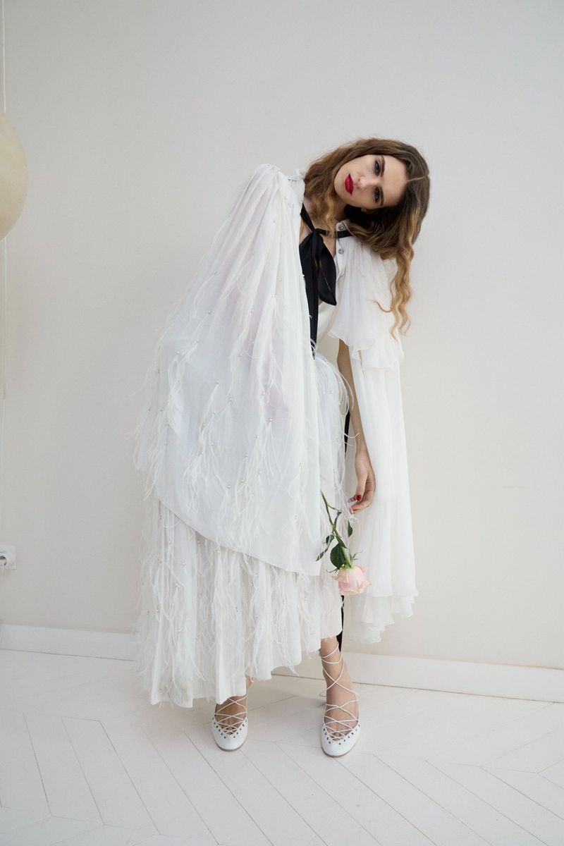 Elegant woman in feathered white dress and cape poses indoors, holding a pink rose, with neutral backdrop. Fashion photography.