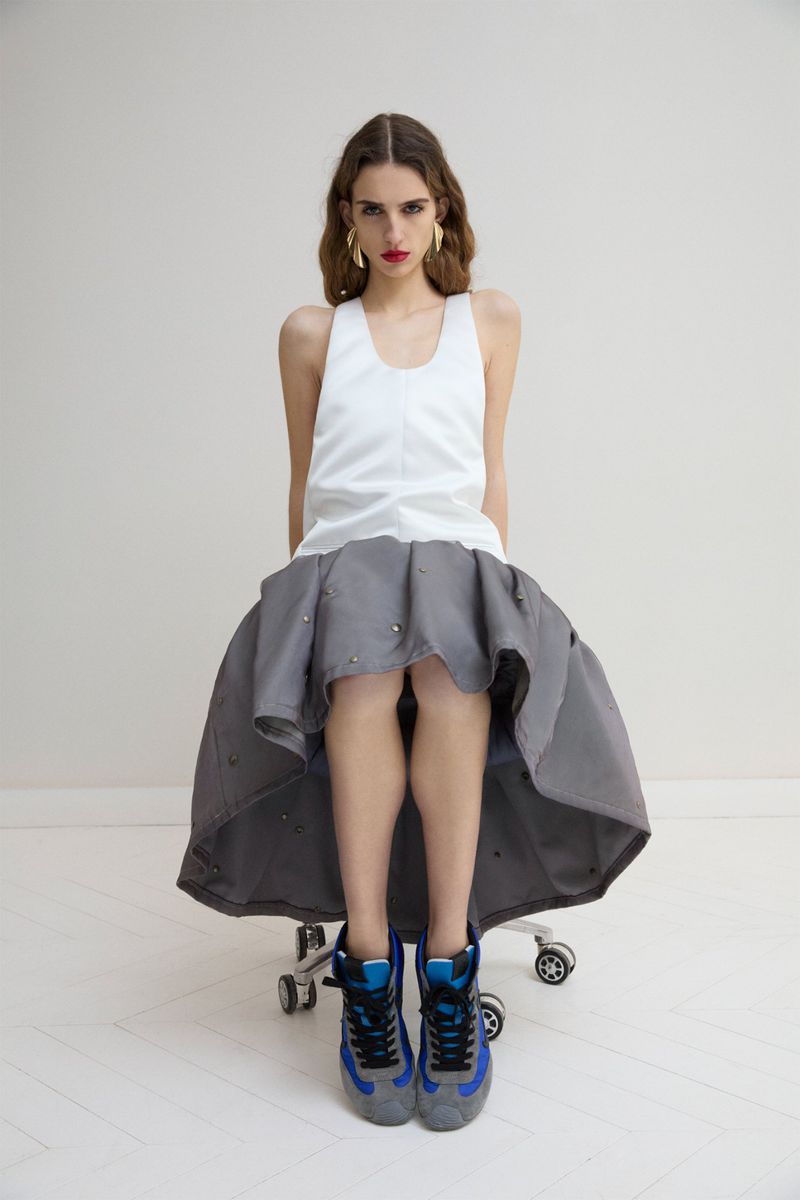 A young woman with long wavy hair sits on a rolling chair wearing a white tank top, full gray skirt, gold earrings, and sneakers in a studio.