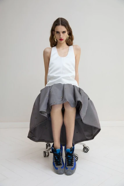 A young woman with long wavy hair sits on a rolling chair wearing a white tank top, full gray skirt, gold earrings, and sneakers in a studio.