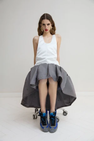 A young woman with long wavy hair sits on a rolling chair wearing a white tank top, full gray skirt, gold earrings, and sneakers in a studio.