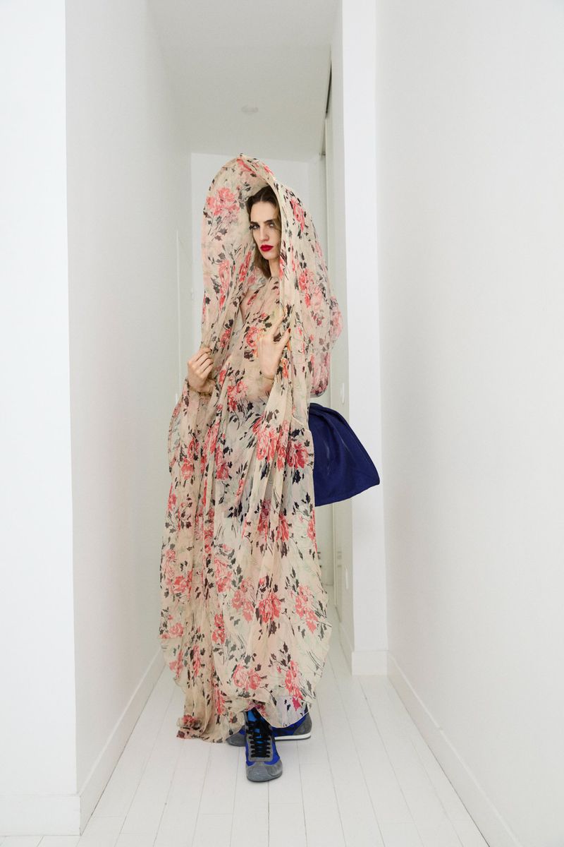 A woman stands in a hallway draped in a semi-transparent floral shawl and a blue dress, with gray sneakers visible under the fabric.