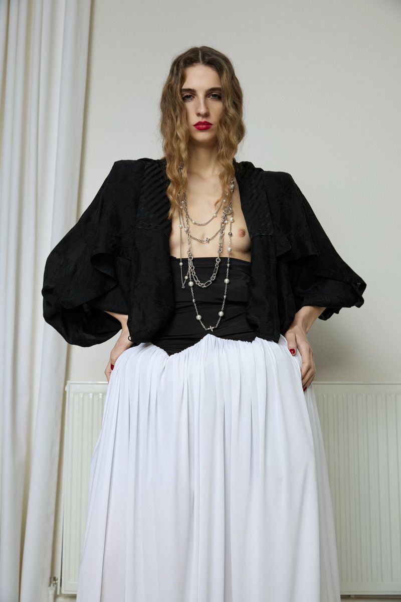 A slender woman with long wavy hair wears a black blouse, silver necklaces, and white skirt against a plain indoor background.