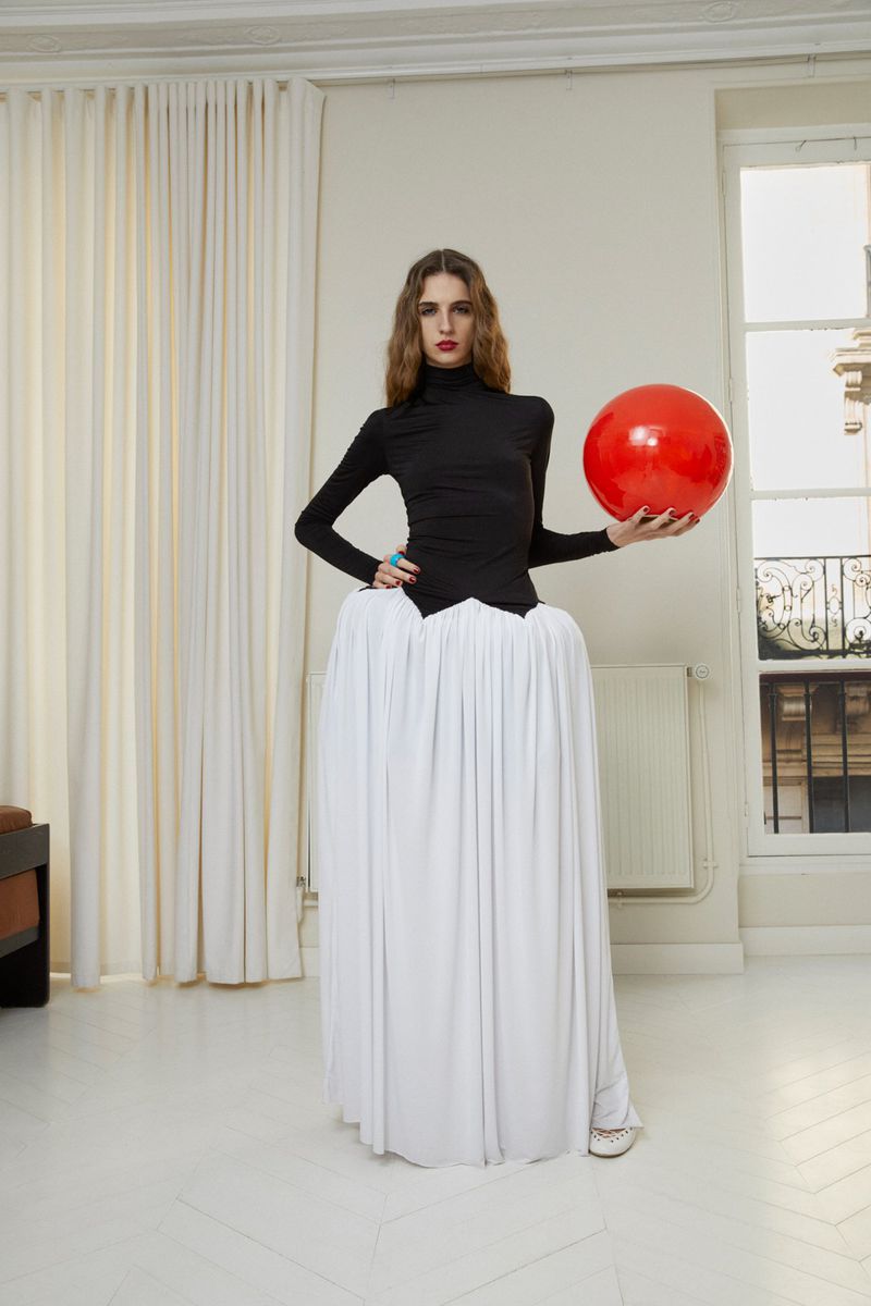 Fashion model poses indoors wearing a black top and white voluminous skirt, holding a red ball against a neutral backdrop.