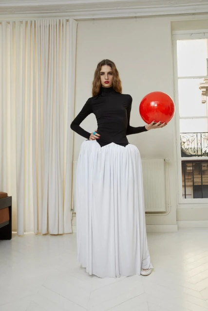 Fashion model poses indoors wearing a black top and white voluminous skirt, holding a red ball against a neutral backdrop.