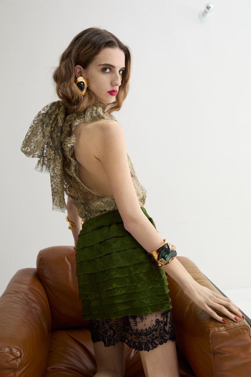 A woman poses in a studio, showcasing a lace top, green skirt, and large earrings. She leans on a leather chair, looking over shoulder.