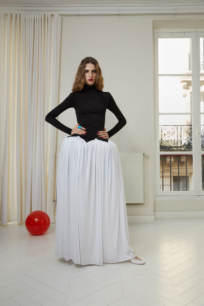Adult woman in a black turtleneck and white flowing skirt poses in a white room with a window and a red balloon, fashion studio shot.