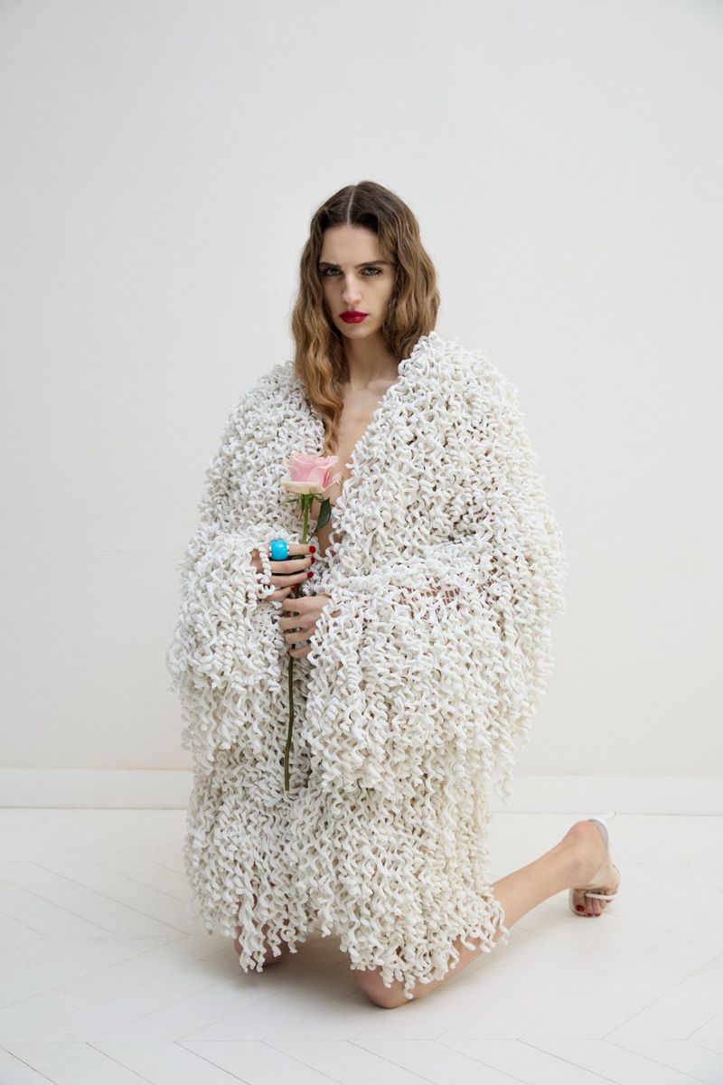 Model kneeling while wearing a textured white knitted cardigan and holding a single pink rose against a white seamless background.