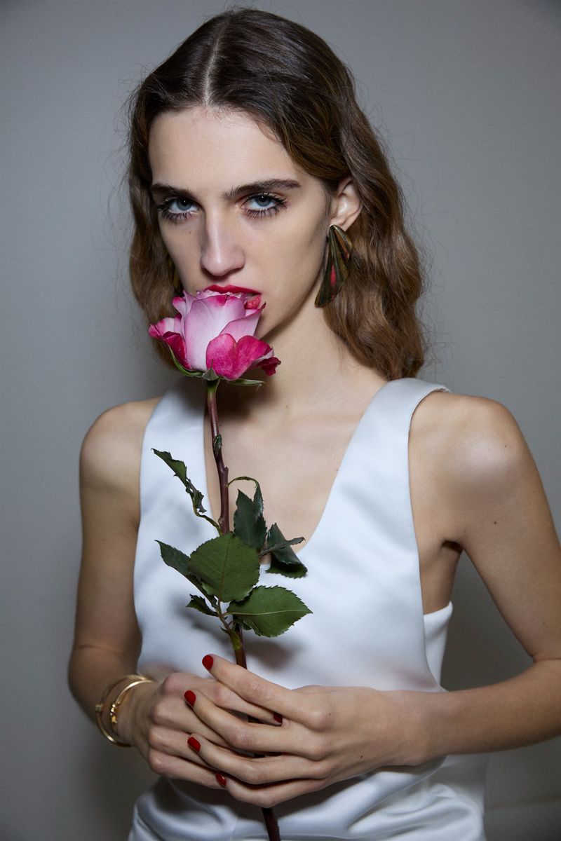 Elegant woman with long hair in white top holds a pink rose to her mouth in a studio, accented by gold bracelets and red nails.