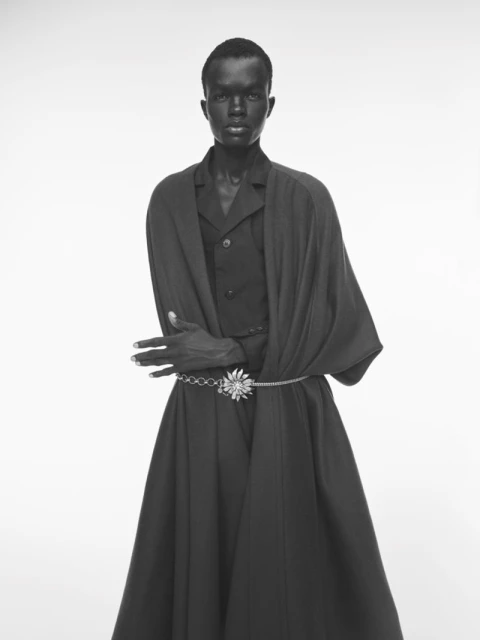 Monochrome portrait of a Black male model in a vest, coat, and long skirt, cinched with a silver belt, posing in a bright studio setting.