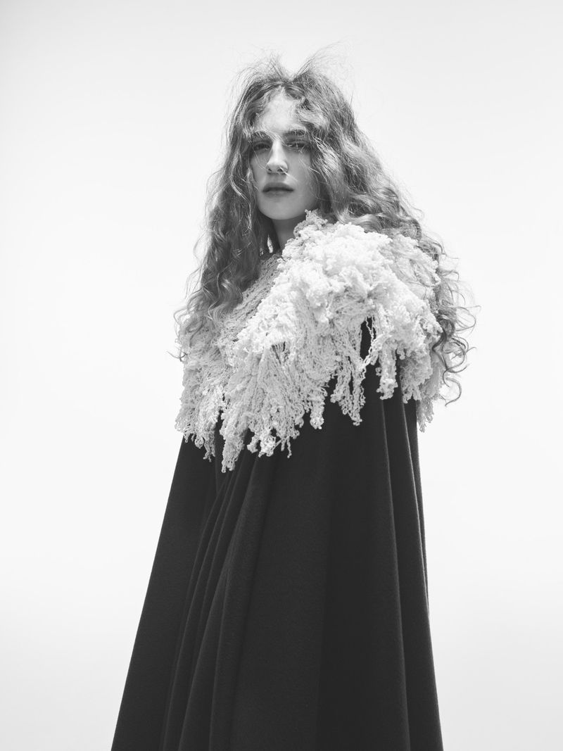 Black and white portrait of a woman wearing a dark cape with a large, white, fringed collar against a bright, neutral background.