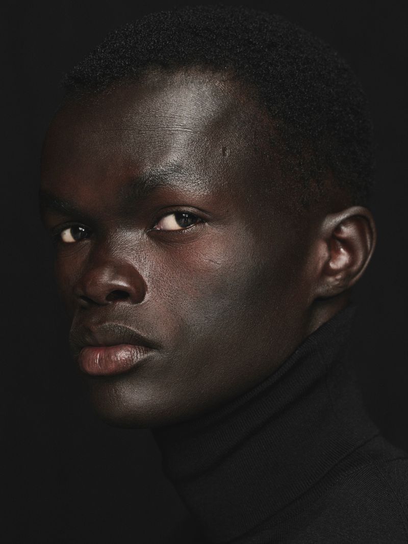 Close-up portrait of a dark-skinned adult male wearing a black turtleneck sweater against a black background, capturing a neutral expression.
