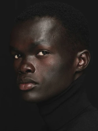 Close-up portrait of a dark-skinned adult male wearing a black turtleneck sweater against a black background, capturing a neutral expression.