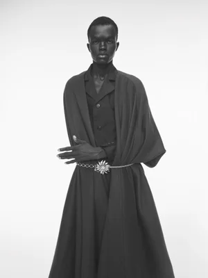 Model wearing a dark collared shirt and long draped coat with a statement belt against a plain white background.