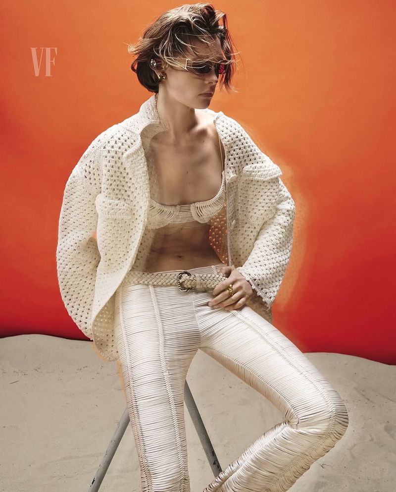 A model poses against an orange background, wearing a woven bra top, open weave jacket, and woven pants while sitting on a stool.