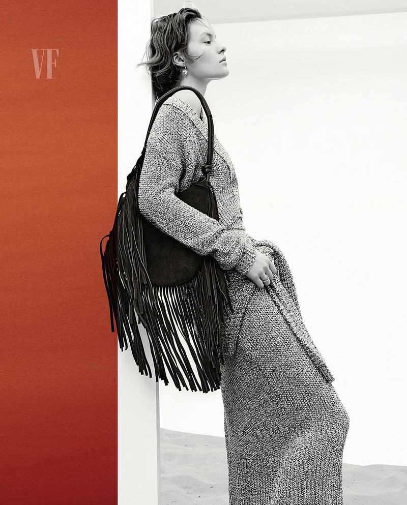 Adult female in a grey knit dress and carrying a black fringe bag, posing near a red wall and white backdrop in a fashion editorial shot.