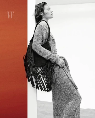 Adult female in a grey knit dress and carrying a black fringe bag, posing near a red wall and white backdrop in a fashion editorial shot.